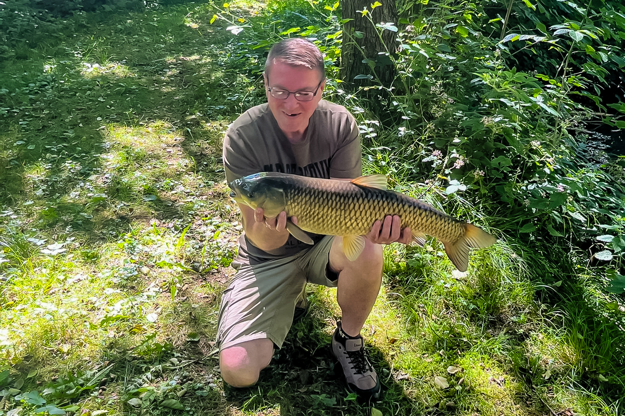 Gastangler kniet im Gras an der Teichanlage und hält einen großen Karpfen